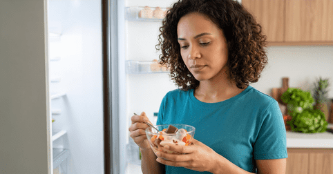 Mulher ansiosa na cozinha à noite olhando para um pedaço de bolo na geladeira, ilustrando como controlar a vontade de comer doce e a compulsão alimentar